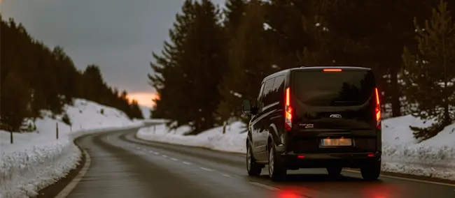 Furgoneta negra circulando por carretera nevada al atardecer. Transporte a estaciones de esquí alternativas.